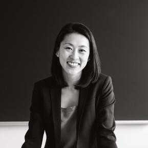 A person is shown standing with their hands on a table, wearing a black blazer over a light-colored top. The image is in black and white, emphasizing the formal setting. The background appears dark, possibly a classroom or conference room.