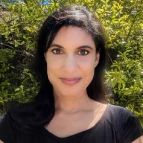 A woman with long black hair and a subtle smile poses in front of bright green foliage, wearing a black shirt and illuminated by natural sunlight.