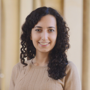 A woman with curly dark hair is smiling at the camera. She is wearing a light brown sweater and is standing in an architectural setting with soft lighting.