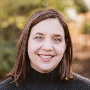 a woman with shoulder-length brown hair and a warm smile is standing outdoors. She is wearing a dark turtleneck sweater. The background is softly blurred with greenery and warm light, suggesting a natural setting.
