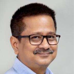 A middle-aged man with short, dark hair and a mustache is smiling at the camera. He is wearing glasses and a light blue collared shirt. The background is a soft, neutral color that keeps the focus on his friendly expression.