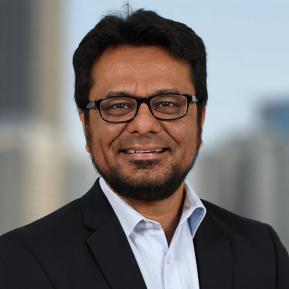 A man with dark hair and a beard is smiling at the camera. He is wearing glasses and a dark suit jacket over a light blue shirt. The background shows a blurred cityscape, suggesting a professional setting.