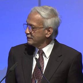 A man with gray hair, wearing glasses and a dark suit, is seen speaking or addressing an audience. He has a slight smile and a thoughtful expression. The background features a blue gradient, adding a professional ambiance to the scene.