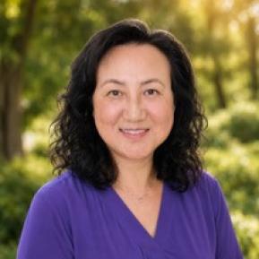 A woman with curly black hair smiles in a green outdoor setting, wearing a purple blouse. Soft sunlight filters through the trees behind her.