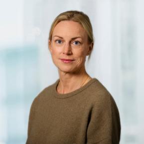 A professional headshot of a woman with straight, blonde hair, wearing a soft, beige sweater. She is facing the camera, smiling gently, with a blurred office background providing natural light.