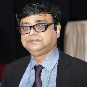 A middle-aged man with wavy black hair and glasses is looking directly at the camera. He is wearing a black suit jacket over a light blue shirt with a collar and a dark red tie. The background is slightly blurred, suggesting an indoor event setting.