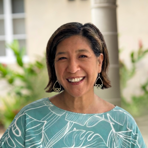 A close-up portrait of a smiling woman with shoulder-length hair. She is wearing a light blue shirt with white leaf patterns and distinctive earrings. The background is softly blurred with greenery and architectural details, suggesting an outdoor setting.