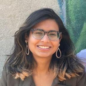 A woman with medium-length, wavy dark hair that has lighter tips is smiling at the camera. She is wearing clear glasses and decorative drop earrings. The background features a textured wall with green hues, and she appears cheerful and friendly.