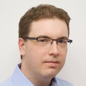 A man with short, wavy brown hair and glasses is looking directly at the camera. He has a neutral expression and is wearing a light blue collared shirt. The background is plain white, which emphasizes his features.