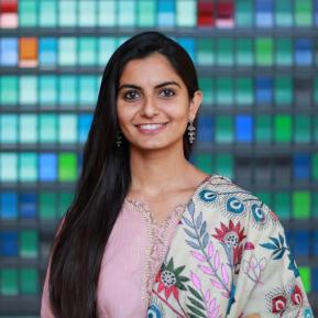 A smiling woman with long dark hair is standing in front of a colorful tiled wall. She is wearing a light pink traditional outfit with intricate embroidery and a decorative shawl featuring floral patterns. 