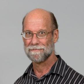 A middle-aged man with thinning hair and a full beard is smiling at the camera. He is wearing glasses and a black and white striped shirt. The background is light gray, allowing his features to stand out clearly.