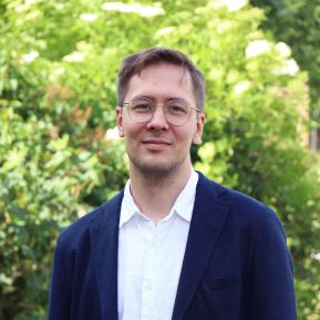 A professional portrait of a man with short brown hair, wearing glasses, a white shirt, and a dark blazer. He is standing outdoors in front of lush green foliage, with a slight smile and a relaxed posture.