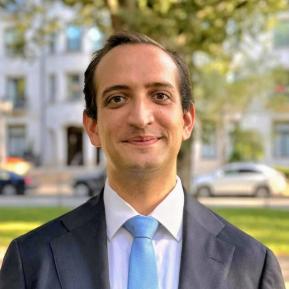 A man with short dark hair and a slight smile stands outdoors. He is wearing a dark suit, a white dress shirt, and a light blue tie. The background features trees and a building.