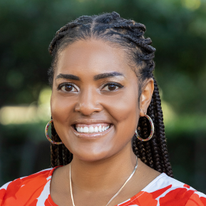 A close-up portrait of a smiling woman with braided hair, wearing colorful earrings and a vibrant red and white top. She appears cheerful, with a natural outdoor background that is softly blurred.