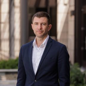A man with short brown hair is standing confidently outdoors. He is wearing a dark suit jacket over a white shirt. The background features elements of modern architecture and greenery, suggesting an urban environment. He has a serious yet approachable expression.