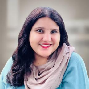 A smiling woman with long, dark hair is wearing a light blue sweater and a soft beige scarf. She is looking at the camera with a friendly expression, showing her red lips. The background is softly blurred, giving focus to her warm demeanor.