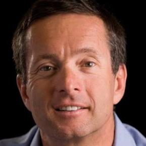 A middle-aged man with short, dark hair and a friendly smile is looking directly at the camera. He is wearing a light blue shirt. The background is dark, which highlights his facial features and creates a professional atmosphere.