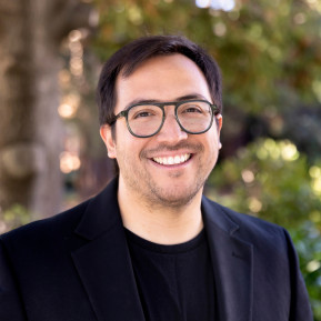 Portrait of a smiling man wearing round glasses and a black blazer outdoors, with trees and greenery blurred in the background.