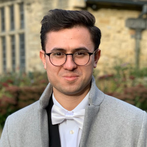 Portrait of a man wearing round glasses, a white bow tie, and a gray overcoat, standing outdoors in front of a stone building and garden.