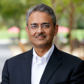 Portrait of a man wearing glasses, a dark suit jacket, and a white shirt, posed outdoors with blurred greenery in the background.