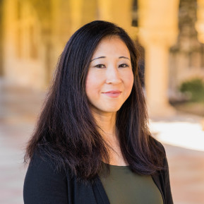 Portrait of a woman with long dark hair wearing a black cardigan and olive-green top, standing beside a column in a sunlit colonnade.