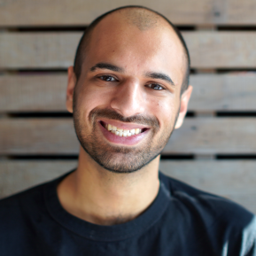 Close-up portrait of a smiling bald man with a short beard, wearing a black shirt against a wooden slat background.