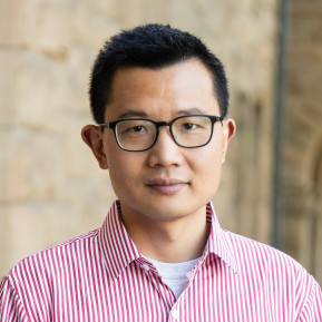 Portrait of a man wearing black glasses and a red-and-white striped button-down shirt, standing in front of a beige stone wall.