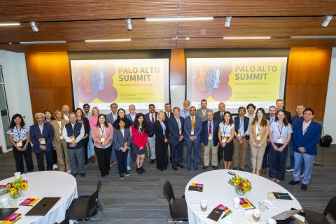 A group of people stands together for a photo at the Palo Alto Summit, which focuses on extending kidney exchange. The event decor features large screens displaying the summit title and theme. In the foreground, there are tables with flowers, coffee cups, and materials from the conference. The attendees are dressed in professional attire and are gathered in a well-lit indoor space.