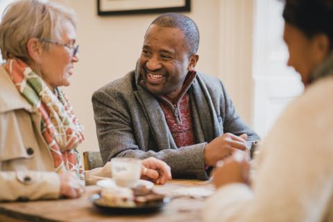 A cozy café scene featuring three individuals seated around a wooden table. One person is wearing a light coat and a colorful scarf, while another is dressed in a gray blazer over a red sweater. In front of them are cups of coffee and a plate of desserts, suggesting a relaxed conversation atmosphere. The background is softly blurred, contributing to the intimate setting.