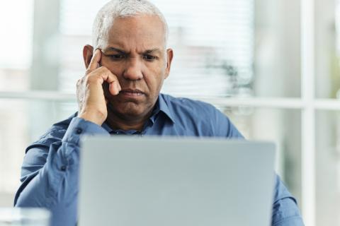 A focused individual is sitting at a desk, resting their chin on their hand while looking at a laptop. They are wearing a blue shirt and are surrounded by a bright, modern office environment with large windows and natural light.