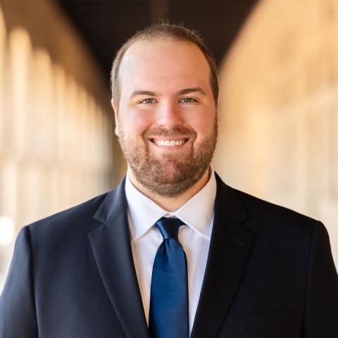 male wearing a blue suit, white shirt and blue tie standing in front of a brown background