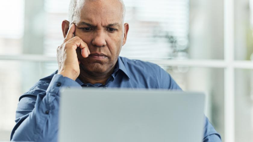 A focused individual is sitting at a desk, resting their chin on their hand while looking at a laptop. They are wearing a blue shirt and are surrounded by a bright, modern office environment with large windows and natural light.