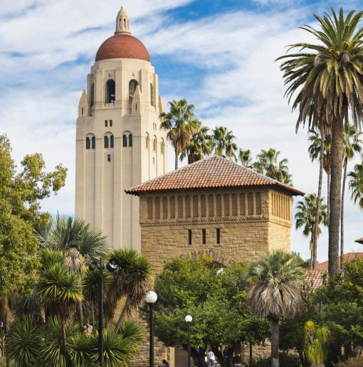 A scenic view of Stanford University featuring the iconic Hoover Tower in the background, surrounded by palm trees and lush greenery. In the foreground, students are sitting on a circular bench and walking along the paths, engaging with their devices and studying. The clouds in the blue sky create a picturesque atmosphere over the campus.
