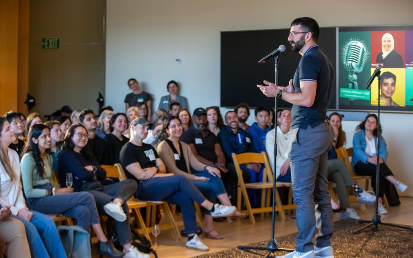 image of male standing in front of mic speaking to a room full of people