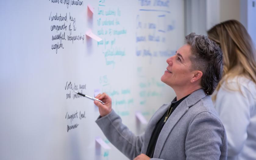 image of female wearing a grey blazer looking at white board with a marker in hand