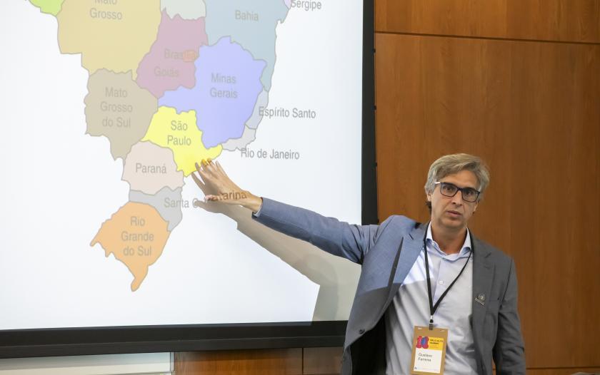 A presenter gestures toward a map of Brazil projected on a screen, highlighting the São Paulo region. The individual is dressed in a blue blazer and light blue shirt, wearing a name tag around their neck. The background features wooden panels and a modern presentation setup, indicating a professional setting.
