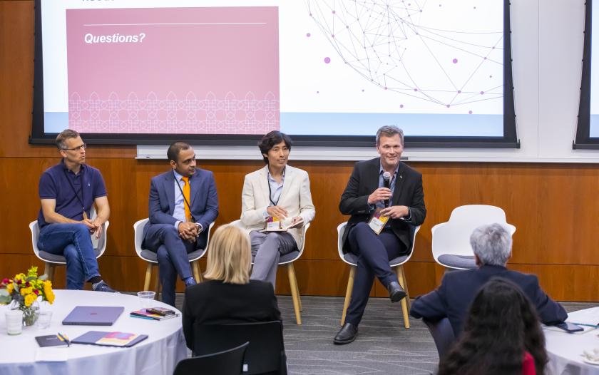 A panel discussion taking place in a conference room. Four individuals are seated on stage, engaged in conversation. In the background, a large screen displays the word "Questions?" along with a network design graphic. A round table with a flower arrangement is visible in the foreground, with people seated around it, some looking towards the panel. The overall setting is professional, with modern furnishings and a relaxed atmosphere.