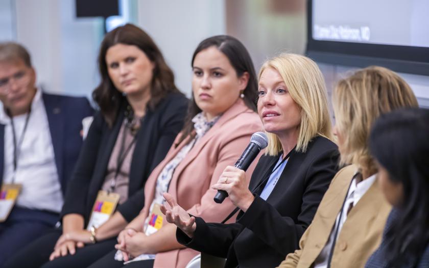 A woman holds a microphone while speaking during a panel discussion, wearing a black blazer over a light blue shirt. She gestures expressively with her hands. Other panelists are seated next to her, dressed in various professional attire. The setting reflects a formal discussion environment, with a presentation screen in the background. The audience can see name tags hanging from the panelists.