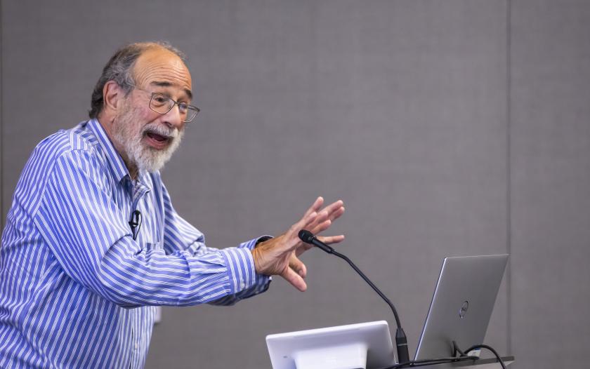 A person in a blue and white striped shirt gestures with both hands while speaking at a podium with a microphone. A laptop is placed on the podium, and the background is a simple gray wall.