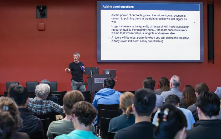 man wearing a black shirt speaking in front of a screen