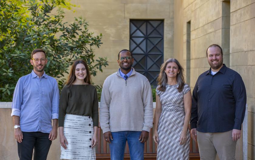 A group of five people poses for a photo outdoors. They stand together, with two individuals on the left, followed by a person in a light sweater at the center, and two more individuals on the right. The background features a stone wall and green foliage. Everyone is dressed in casual to semi-formal attire. Faces are blurred for privacy.