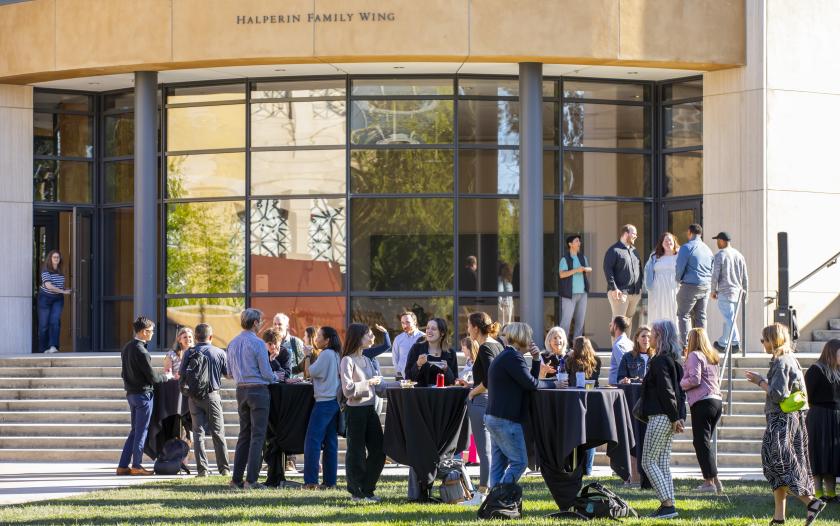 A lively outdoor gathering in front of a building labeled "Halperin Family Wing." Groups of people are mingling near tables, some holding drinks and plates of food. A few attendees are engaged in conversation while others are moving in and out of the building. The architecture features large glass windows that reflect the surroundings, enhancing the modern atmosphere. The setting suggests an informal event or social gathering, with a sunny day contributing to the cheerful ambiance.