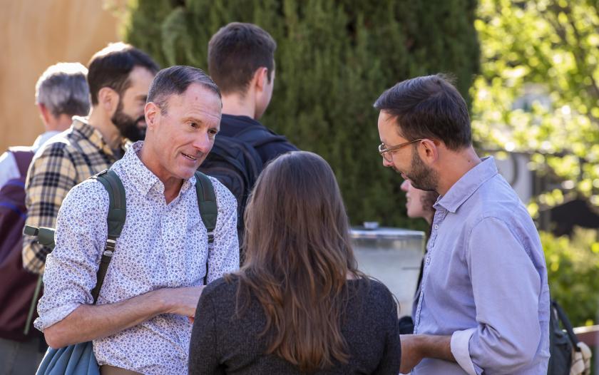 A small group of individuals is engaged in conversation outdoors. One person, wearing a floral-patterned shirt with a backpack, appears to be speaking. A woman with long hair is listening intently. Another individual in a light-colored shirt is also part of the interaction. The background features greenery and hints of a casual, outdoor environment, suggesting a relaxed gathering or networking opportunity.