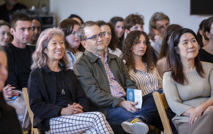 A group of audience members attentively watching a presentation. Some are sitting with arms crossed or holding items in their hands. The background features various people, and the atmosphere appears engaged and focused on the speaker. Each individual is in casual attire, and the setting feels informal yet attentive.