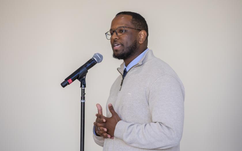 A person stands beside a microphone, speaking while clasping their hands in front. They wear a light sweater and a collared shirt. The background is plain, emphasizing the speaker's engaged demeanor during the presentation.
