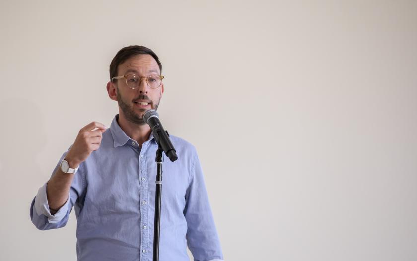 A person stands at a microphone, gesturing with one hand while speaking. They wear a light blue shirt and glasses. The background is plain, and the focus is on the speaker, who appears engaged in delivering a message to an unseen audience.