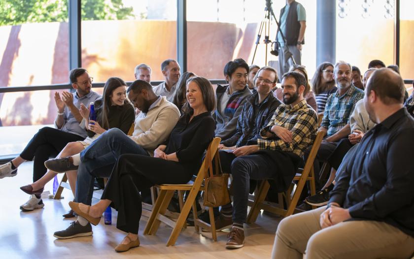 A group of people sit in a well-lit room, facing a presentation, some clapping, while others listen intently. The audience is casually dressed and appears engaged. There’s natural light streaming in through large windows, adding to the inviting atmosphere. A person is seen in the background, seemingly recording or monitoring the event.