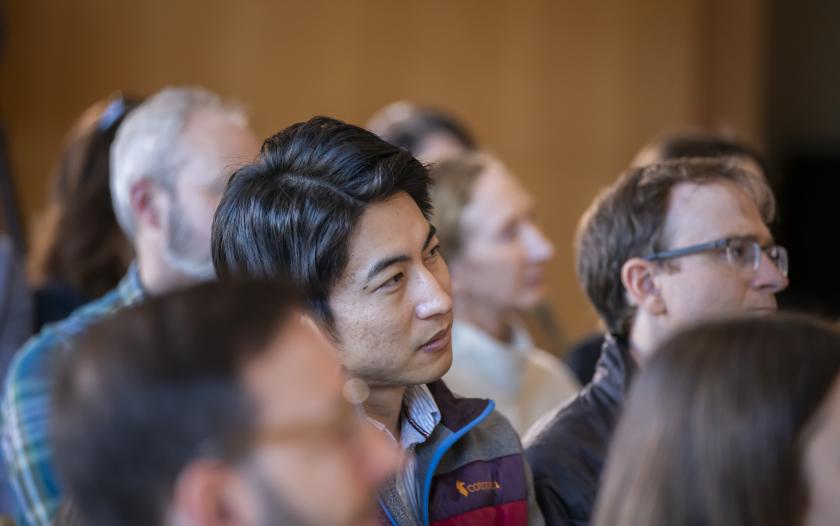 A close-up view of several audience members, primarily focusing on their heads and shoulders. They're engaged in an event, with hair and clothing visible. The background is blurred, emphasizing the expressions and attention of the individuals in the foreground. The atmosphere appears focused, suggesting an attentive audience during a presentation or discussion.