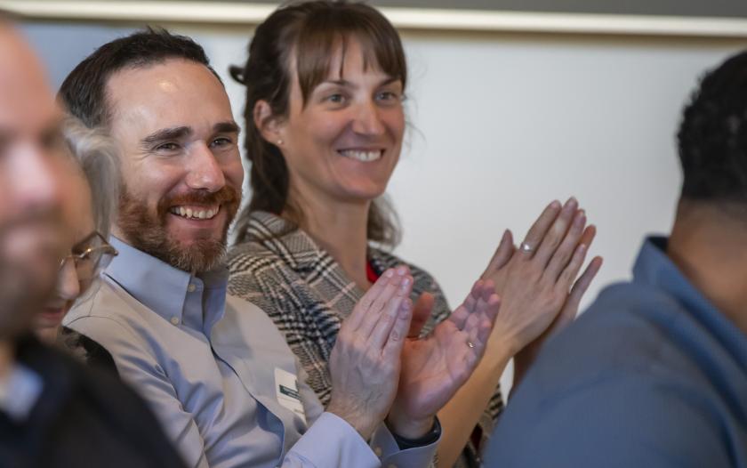 A close-up of two individuals applauding enthusiastically as part of an audience. The person on the left appears to be wearing a light-colored shirt, while the person on the right has a patterned jacket. The focus on their hands and expressions conveys a sense of appreciation and engagement with the event happening in front of them. The background is blurred, highlighting their participation.
