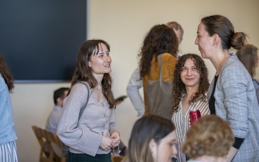A group of individuals is engaged in conversation in a casual indoor setting. One woman is wearing a light-colored sweater and green pants, while another has curly hair and is holding a can of soda. Nearby, a third woman in a patterned jacket is interacting with them. The atmosphere appears friendly and relaxed, suggesting a social or networking event. The background is softly blurred, keeping the focus on the group’s interaction.